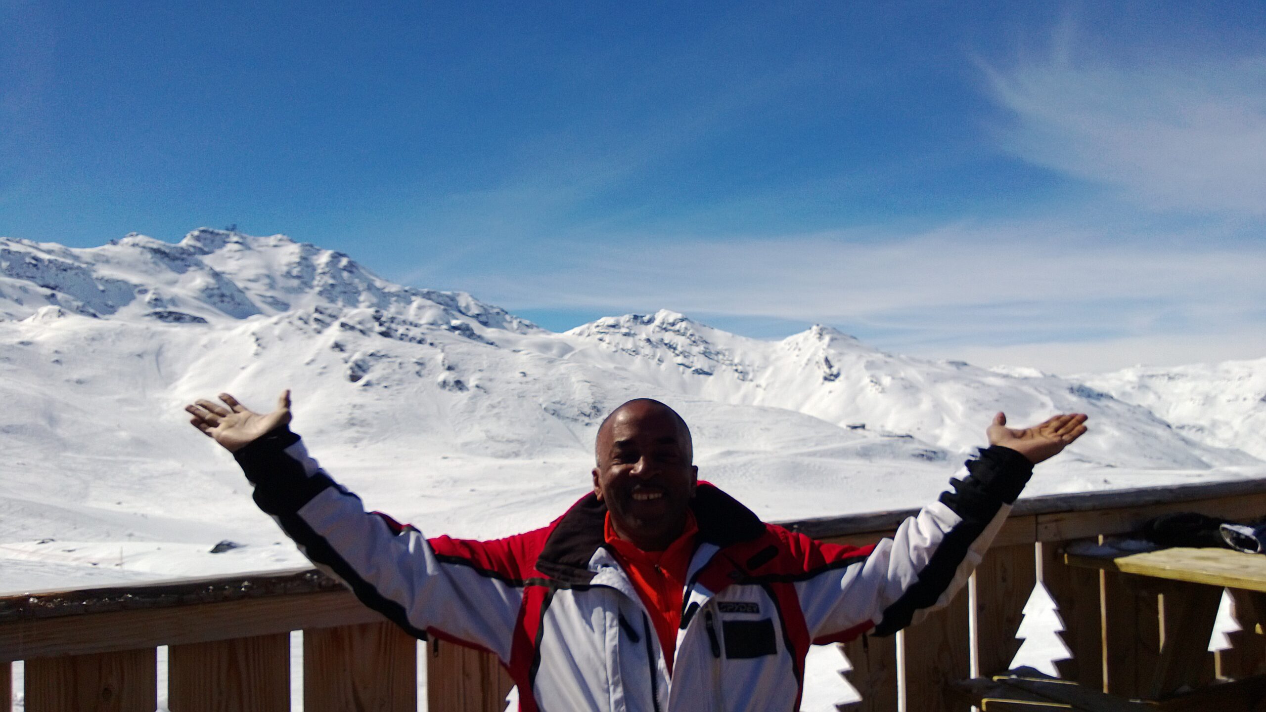 Roy raises his arms in a snowy alpine setting with blue sky behind him.