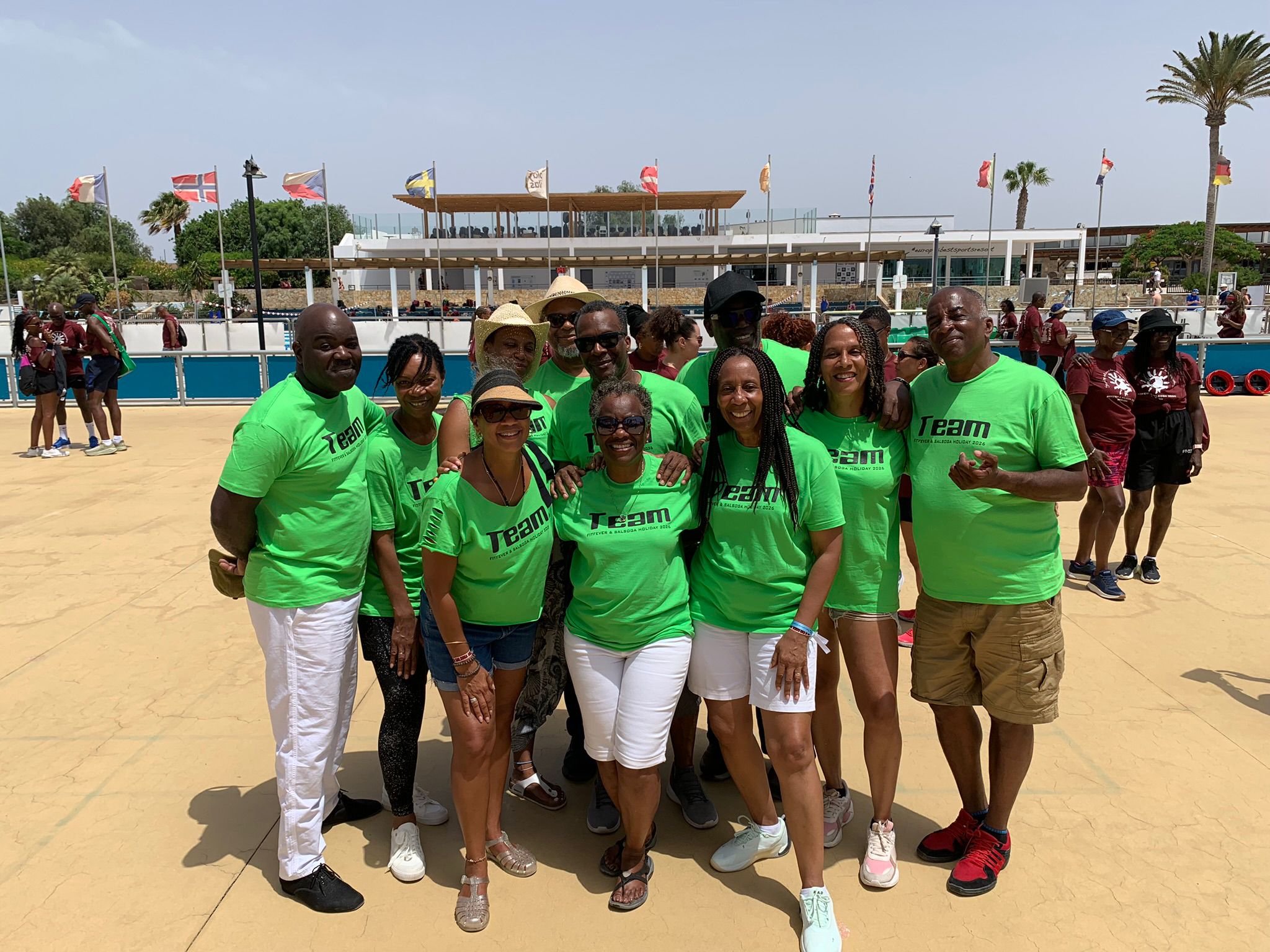 Group of smiling people in matching bright green shirts posing outdoors at a sunny sports venue with flags in the background.